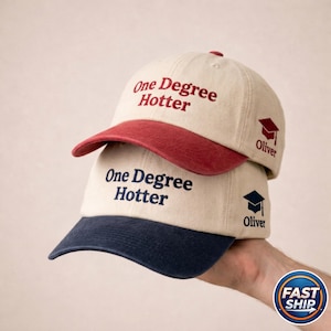 Peut inclure: Deux casquettes de baseball superposées. La casquette supérieure a une visière rouge et le texte "One Degree Hotter" brodé en rouge. La casquette inférieure a une visière bleu marine avec le même texte en bleu marine. Les deux casquettes ont un motif de chapeau de graduation et le nom "Oliver".