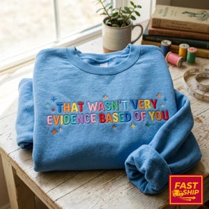 Pode incluir: Uma camisola de gola redonda azul-clara com o texto bordado em várias cores "THAT WASN'T VERY EVIDENCE BASED OF YOU" e pequenos detalhes de estrelas bordadas em cores variadas à volta das letras na parte da frente.