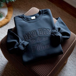 May include: A navy blue sweatshirt with the embroidered text "BAD DAY To be an Alani." The sweatshirt is folded on a brown patterned ottoman. The sleeves are folded, and the overall look is casual and comfortable.