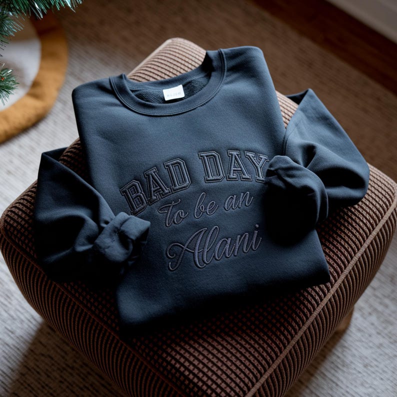 May include: A navy blue sweatshirt with the embroidered text "BAD DAY To be an Alani." The sweatshirt is folded on a brown patterned ottoman. The sleeves are folded, and the overall look is casual and comfortable.