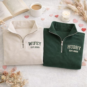 May include: Two matching cream and green quarter-zip sweatshirts. The cream sweatshirt has the word "Wifey" and "EST.2026" embroidered in green. The green sweatshirt has the word "Hubby" and "EST.2026" embroidered in white. The sweatshirts are on a white surface.