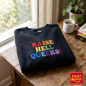 May include: A black crewneck sweatshirt featuring embroidered text that reads RAISE HELL QUEERS! in a rainbow color palette. The word RAISE is in red and orange, HELL is in yellow and green, and QUEERS! is in blue, purple, and pink.