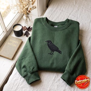 May include: A forest green crewneck sweatshirt featuring a detailed black embroidered raven centred on the chest. The garment has long sleeves with ribbed cuffs and a ribbed neckline, showcasing a clean, solid-coloured fabric design.