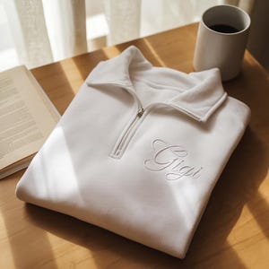 Könnte beinhalten: Ein gefalteter weißer Pullover mit einem Reißverschlusskragen und dem gestickten Namen "Gigi" in Schreibschrift. Der Pullover liegt auf einer Holzoberfläche, neben einer weißen Tasse und einem offenen Buch.