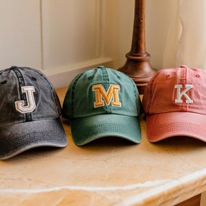 May include: Three baseball caps with embroidered letters. The first cap is a faded black with a white "J". The second is green with a gold "M". The third is a dusty rose color with a white "K".