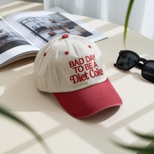 Peut inclure: Casquette de baseball crème avec une visière et un bouton supérieur rouges. Le chapeau porte l'inscription brodée en rouge "BAD DAY TO BE A Diet Coke". Des lunettes de soleil et un magazine sont en arrière-plan.