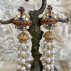 May include: A pair of ornate earrings with a gold-tone setting. The earrings feature a cross design with red and blue accents, surrounded by clear and red stones. Below, dangling strands of white pearls cascade from a gold filigree bell shape.