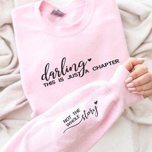 以下が含まれることがあります： 「darling, this is just a chapter」の黒い文字がプリントされた、明るいピンク色のスウェットシャツ。袖には「not the whole story」の文字。柔らかい素材のスウェットシャツです。