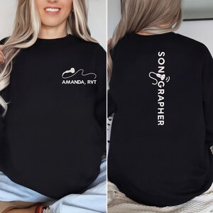 Peut inclure: Sweat-shirt noir avec texte et graphiques blancs. L'avant présente une image d'échographie stylisée et le texte "AMANDA, RVT". Le dos affiche le mot "SONOGRAPHER" verticalement.