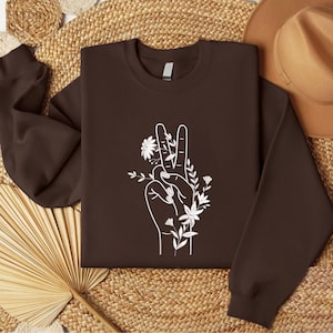 May include: A brown sweatshirt featuring a white graphic of a hand making the peace sign, adorned with floral elements. The design is centered on the front of the long-sleeved crew neck top. The sweatshirt is displayed on a woven surface.