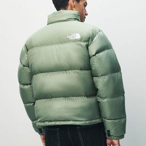 Puede incluir: Una chaqueta acolchada verde salvia con cuello alto y el nombre de la marca impreso en blanco en la parte superior de la espalda. La chaqueta se lleva sobre unos vaqueros oscuros. La chaqueta tiene un dise&ntilde;o acolchado.