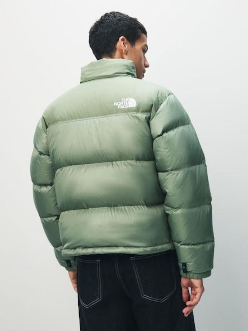 Puede incluir: Una chaqueta acolchada verde salvia con cuello alto y el nombre de la marca impreso en blanco en la parte superior de la espalda. La chaqueta se lleva sobre unos vaqueros oscuros. La chaqueta tiene un dise&ntilde;o acolchado.