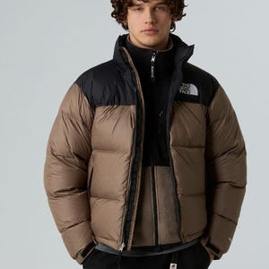 Puede incluir: Chaqueta acolchada marr&oacute;n y negra con cuello alto y cierre de cremallera. El logotipo de la marca est&aacute; en el pecho. El modelo lleva una chaqueta polar negra debajo.