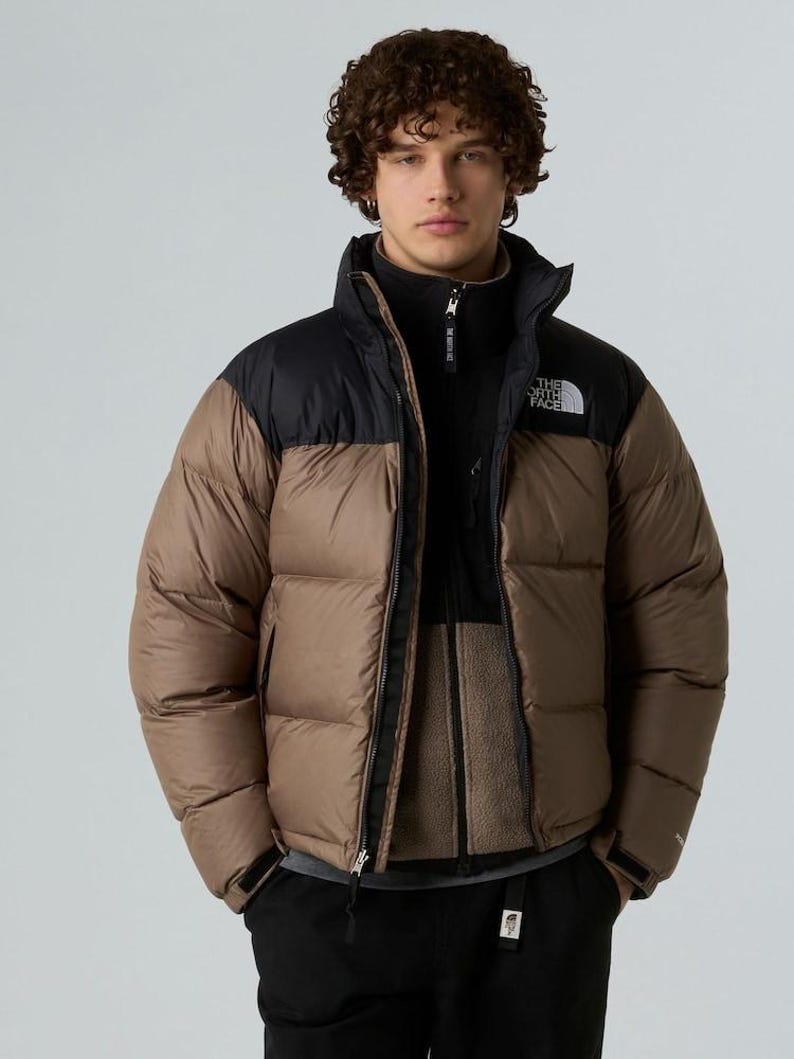Puede incluir: Chaqueta acolchada marr&oacute;n y negra con cuello alto y cierre de cremallera. El logotipo de la marca est&aacute; en el pecho. El modelo lleva una chaqueta polar negra debajo.