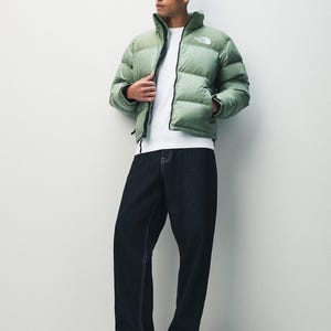 Puede incluir: Una chaqueta acolchada verde claro con cremallera negra y el logotipo de la marca en el pecho. La chaqueta se combina con una camiseta blanca, vaqueros azul oscuro y zapatillas negras. El conjunto es informal y elegante.