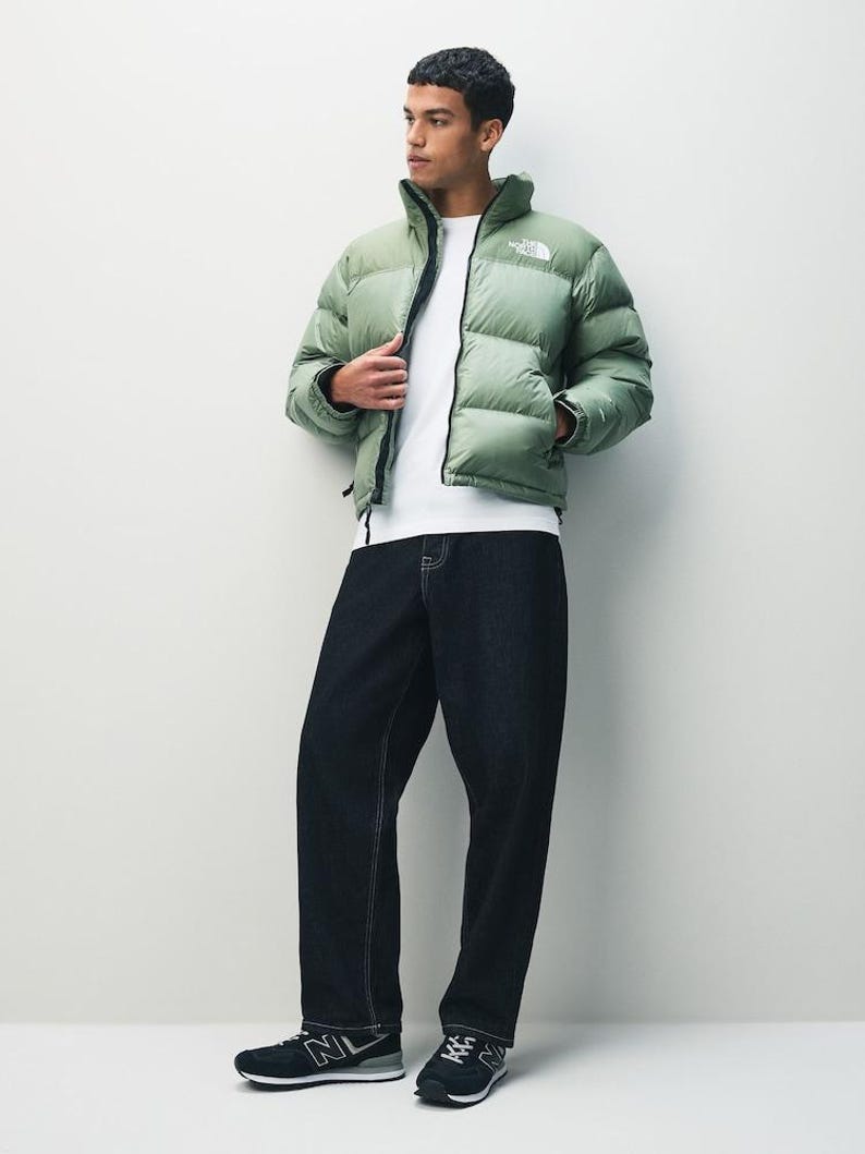 Puede incluir: Una chaqueta acolchada verde claro con cremallera negra y el logotipo de la marca en el pecho. La chaqueta se combina con una camiseta blanca, vaqueros azul oscuro y zapatillas negras. El conjunto es informal y elegante.