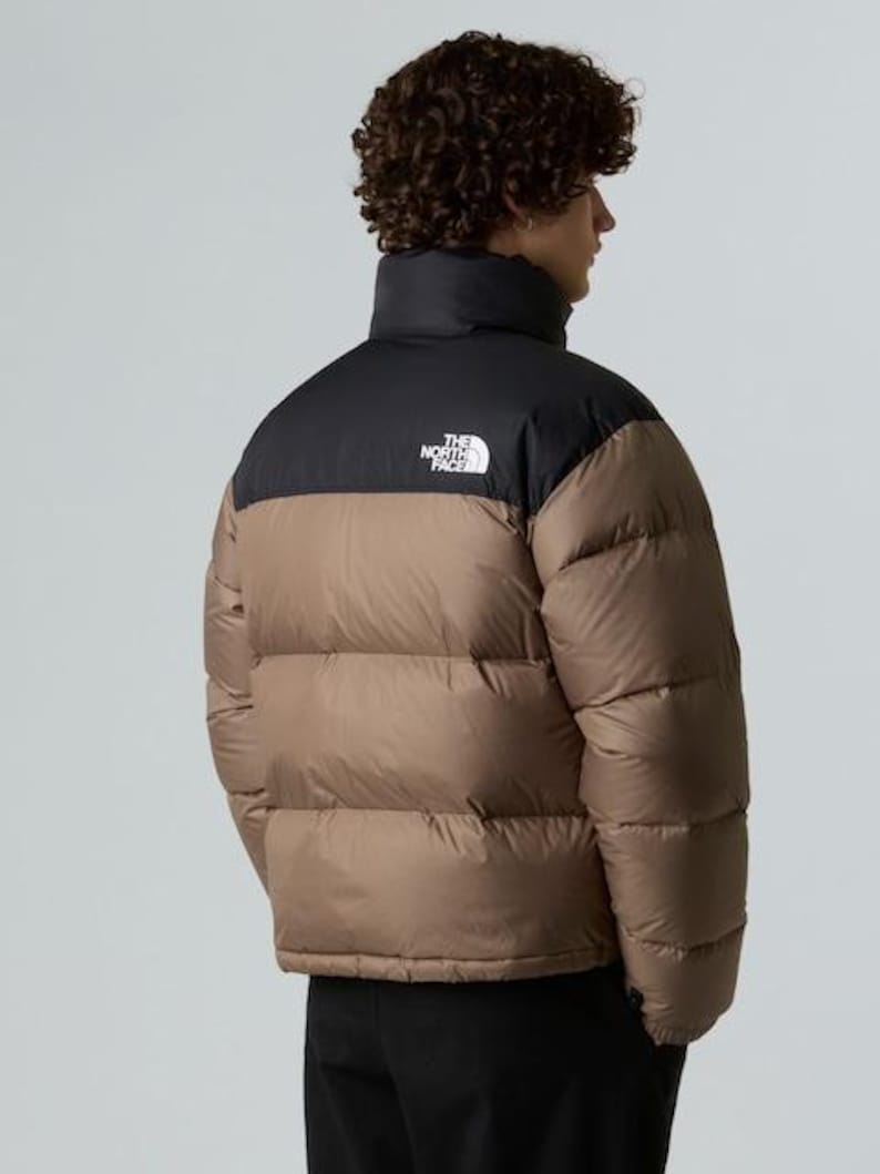 Puede incluir: Chaqueta acolchada marr&oacute;n y negra con cuello alto. La chaqueta tiene una secci&oacute;n superior negra y una secci&oacute;n inferior marr&oacute;n. El logotipo de The North Face es visible en la parte superior izquierda del pecho. La chaqueta la lleva una persona.