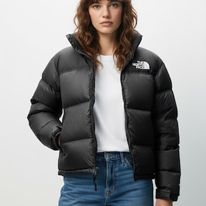 Puede incluir: Chaqueta acolchada negra con cuello alto y logotipo blanco en la parte superior izquierda del pecho. La chaqueta est&aacute; abrochada y se lleva sobre una camiseta blanca y unos vaqueros azules. El logotipo de The North Face es visible.