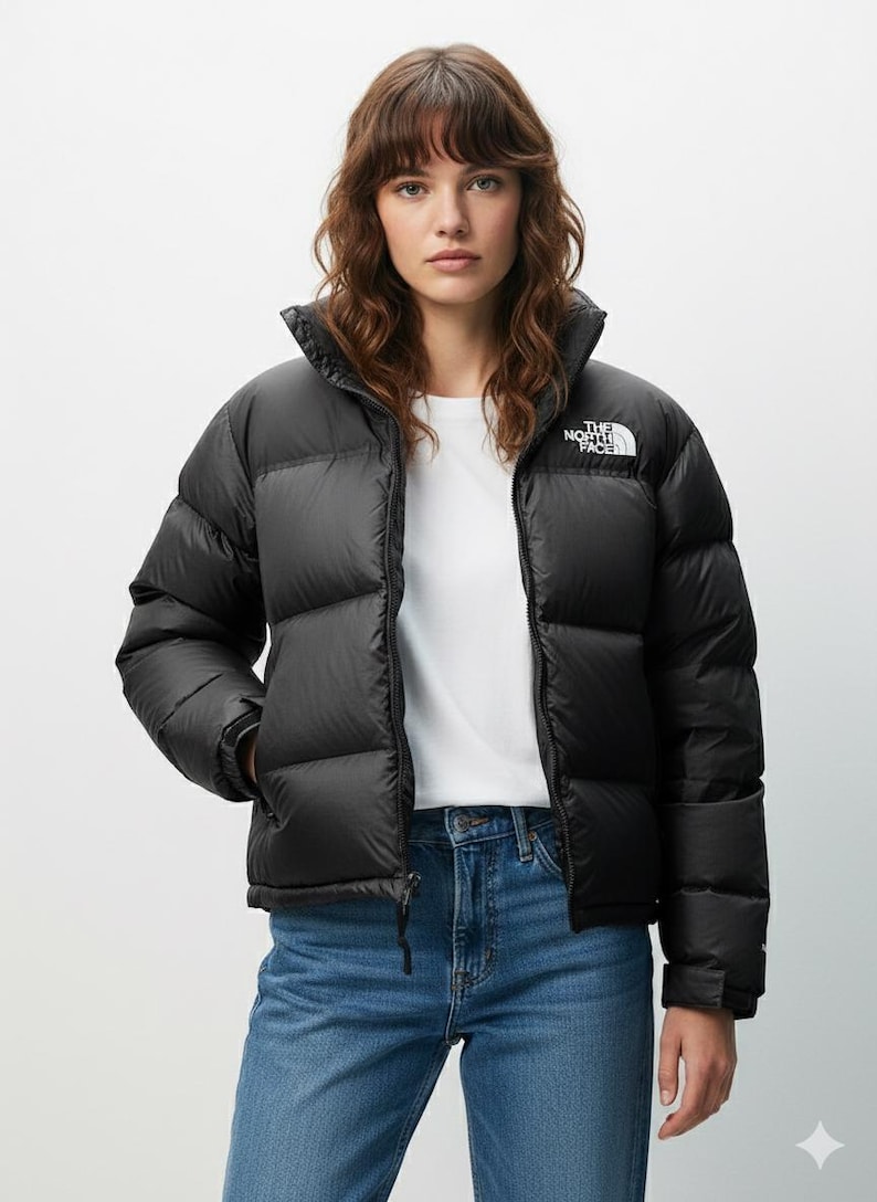 Puede incluir: Chaqueta acolchada negra con cuello alto y logotipo blanco en la parte superior izquierda del pecho. La chaqueta est&aacute; abrochada y se lleva sobre una camiseta blanca y unos vaqueros azules. El logotipo de The North Face es visible.