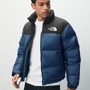 Puede incluir: Chaqueta acolchada azul y negra con el texto "THE NORTH FACE" en el pecho. La chaqueta tiene un cuello alto y cierre de cremallera. El modelo lleva una camiseta blanca debajo.