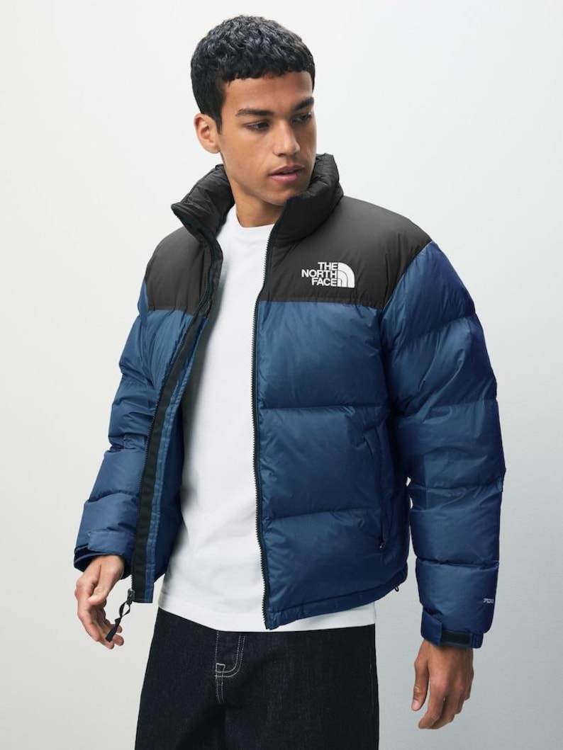 Puede incluir: Chaqueta acolchada azul y negra con el texto "THE NORTH FACE" en el pecho. La chaqueta tiene un cuello alto y cierre de cremallera. El modelo lleva una camiseta blanca debajo.