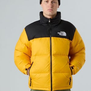 Puede incluir: Chaqueta acolchada amarilla y negra con gorro negro. La chaqueta tiene cuello alto y cremallera frontal. El logo de The North Face est&aacute; en el pecho izquierdo. El modelo lleva pantalones verdes.