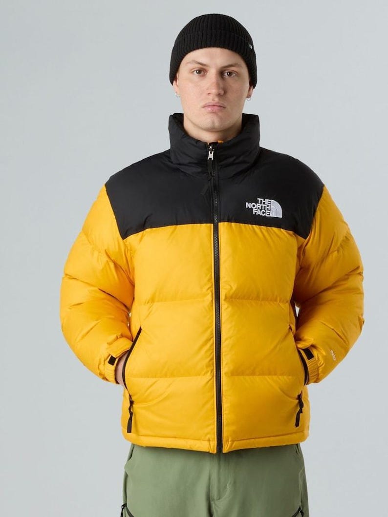Puede incluir: Chaqueta acolchada amarilla y negra con gorro negro. La chaqueta tiene cuello alto y cremallera frontal. El logo de The North Face est&aacute; en el pecho izquierdo. El modelo lleva pantalones verdes.