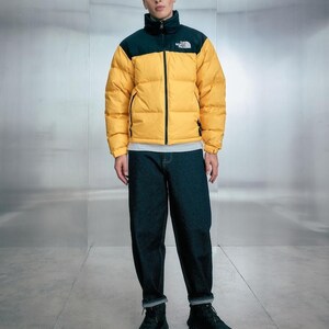 Puede incluir: Una persona con una chaqueta acolchada amarilla y negra con el texto "The North Face" en el pecho. La chaqueta se combina con vaqueros azul oscuro y botas negras. La chaqueta tiene un cuello alto y cierre de cremallera.