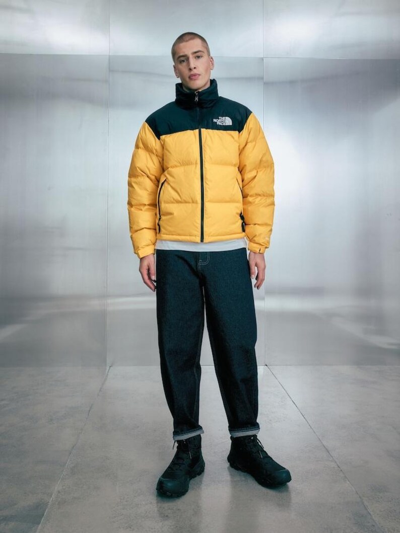 Puede incluir: Una persona con una chaqueta acolchada amarilla y negra con el texto "The North Face" en el pecho. La chaqueta se combina con vaqueros azul oscuro y botas negras. La chaqueta tiene un cuello alto y cierre de cremallera.