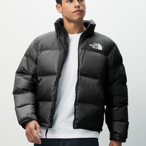 May include: Black puffer jacket with a high collar and a white logo on the chest. The jacket is fully zipped and has a quilted design. The model is wearing a white t-shirt underneath. The North Face brand.