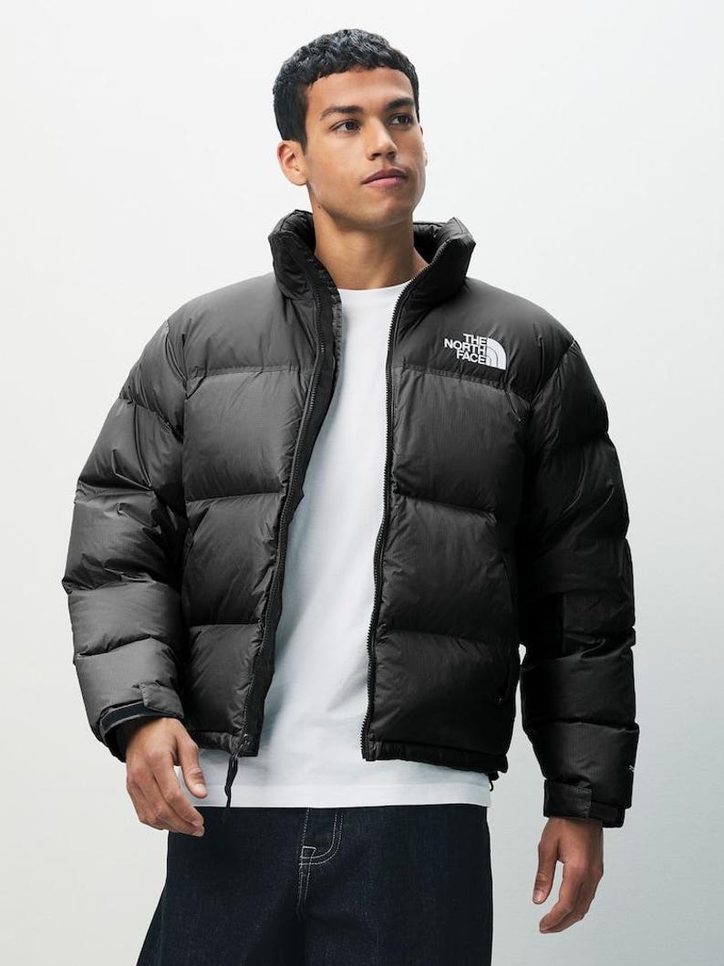 Puede incluir: Chaqueta acolchada negra con cuello alto y logotipo blanco en el pecho. La chaqueta est&aacute; completamente cerrada con cremallera y tiene un dise&ntilde;o acolchado. El modelo lleva una camiseta blanca debajo. Marca The North Face.