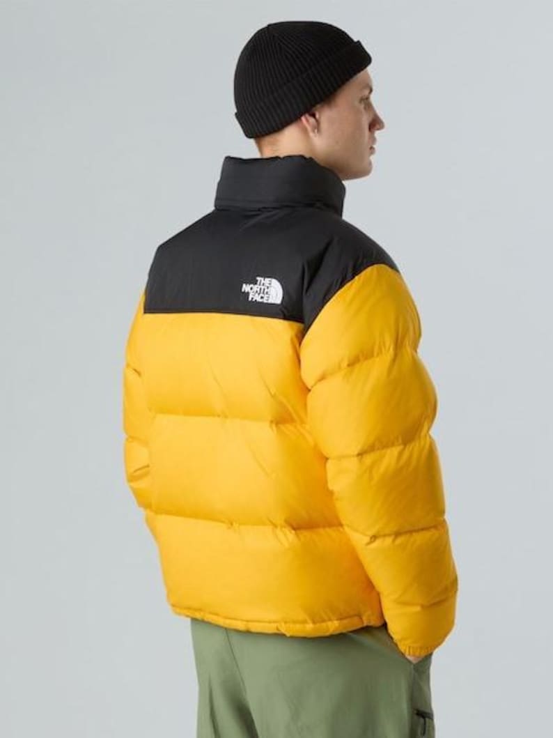 Puede incluir: Un gorro negro y una chaqueta acolchada amarilla y negra. La chaqueta tiene un cuello alto y el logotipo de la marca en el pecho. El modelo lleva pantalones verdes. La chaqueta es una prenda de abrigo c&aacute;lida e informal.