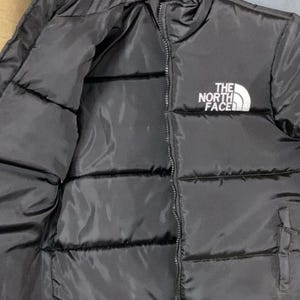 Puede incluir: Chaqueta acolchada negra con cremallera completa y el logotipo blanco "THE NORTH FACE" en la parte superior izquierda del pecho. La chaqueta es acolchada y parece estar hecha de un material liso e impermeable.
