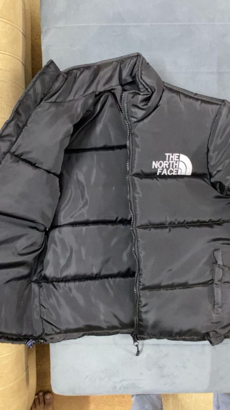 Puede incluir: Chaqueta acolchada negra con cremallera completa y el logotipo blanco "THE NORTH FACE" en la parte superior izquierda del pecho. La chaqueta es acolchada y parece estar hecha de un material liso e impermeable.