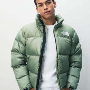 Puede incluir: Chaqueta acolchada verde salvia con cuello alto y cremallera negra. La chaqueta presenta el logotipo blanco "THE NORTH FACE" en el pecho. La persona lleva una camiseta blanca y vaqueros azul oscuro.