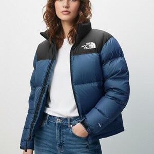 Puede incluir: Chaqueta acolchada azul y negra con cuello alto y cierre de cremallera. El logotipo de la marca est&aacute; en el pecho. La modelo lleva vaqueros azules y una camiseta blanca.