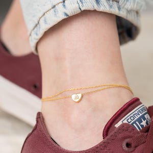 May include: A delicate gold anklet with a heart-shaped charm. The anklet is worn on an ankle, with a burgundy shoe and denim jeans in the background. The heart charm has a small design in the center.