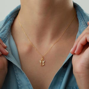 Op de afbeelding: Een goudkleurige ketting met een hanger in de vorm van een engel. De engel hanger heeft kleine, heldere edelstenen op de vleugels en het lichaam. De ketting wordt gedragen tegen een lichtblauw spijkergoed overhemd.