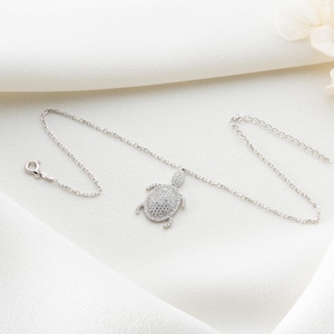 Può includere: Una collana color argento con un ciondolo a forma di tartaruga. Il ciondolo è ricoperto di piccole pietre scintillanti. La collana è esposta su una superficie bianca e strutturata. La collana ha una chiusura a fermaglio.