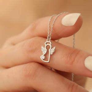 Op de afbeelding: Een zilveren engel hanger ketting. De hanger heeft een engel omtrek met kristallen accenten op de vleugels en het lichaam. De ketting wordt vastgehouden door een hand met verzorgde nagels.