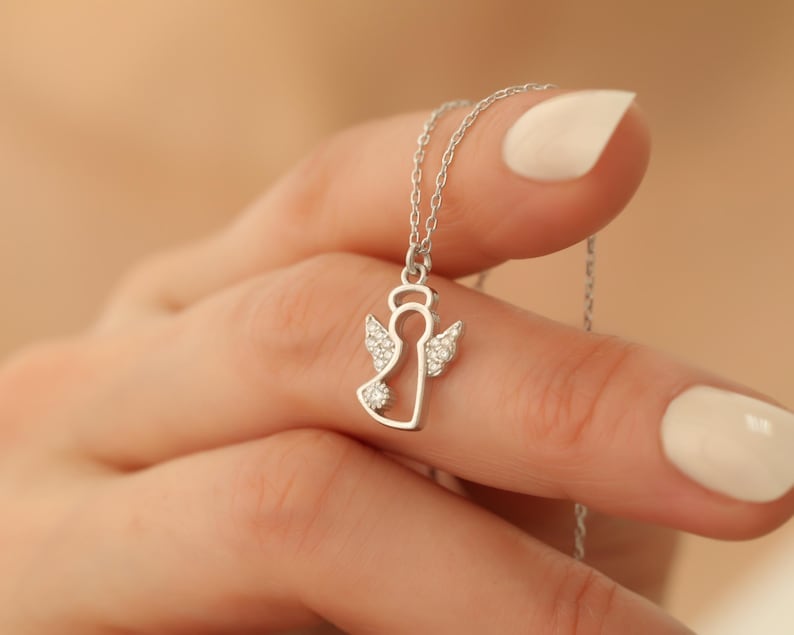 Op de afbeelding: Een zilveren engel hanger ketting. De hanger heeft een engel omtrek met kristallen accenten op de vleugels en het lichaam. De ketting wordt vastgehouden door een hand met verzorgde nagels.