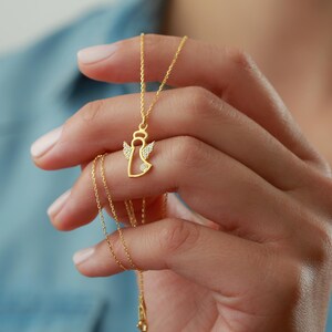 Op de afbeelding: Een gouden engel hanger ketting. De engel heeft een halo en vleugels versierd met kleine, heldere edelstenen. De ketting is fijn en goudkleurig. De hanger wordt in een hand gehouden tegen een blauwe achtergrond.