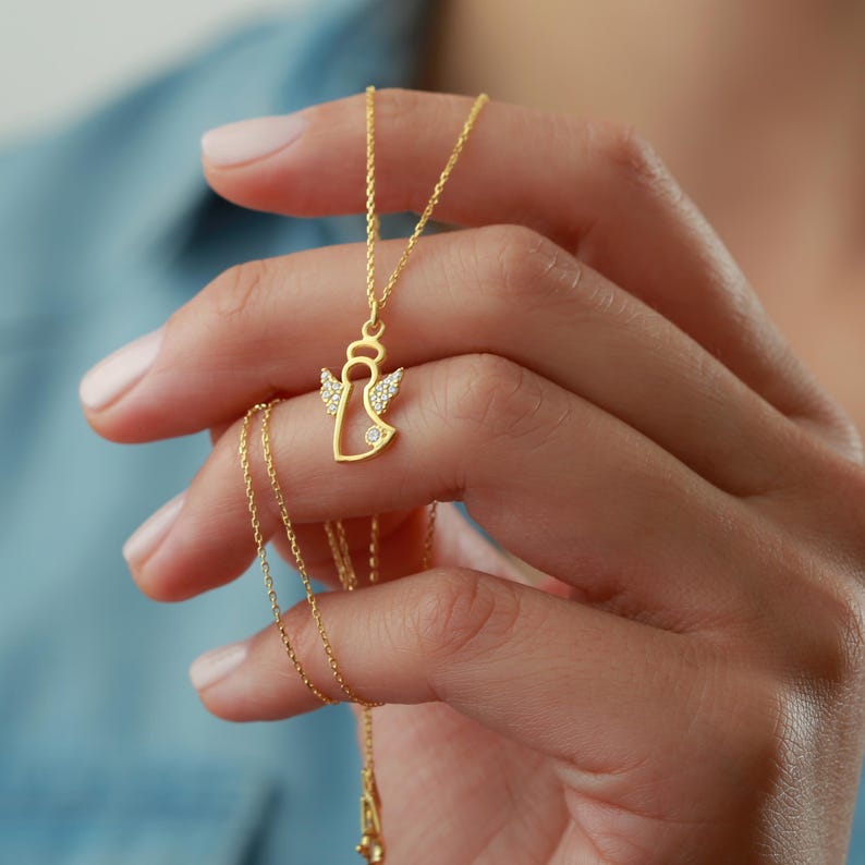 Op de afbeelding: Een gouden engel hanger ketting. De engel heeft een halo en vleugels versierd met kleine, heldere edelstenen. De ketting is fijn en goudkleurig. De hanger wordt in een hand gehouden tegen een blauwe achtergrond.