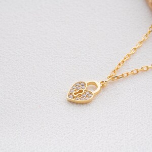 May include: A gold-colored necklace featuring a heart-shaped pendant designed as a padlock. The pendant is encrusted with small, clear gemstones. The necklace chain is delicate and also gold-colored, resting on a white surface.