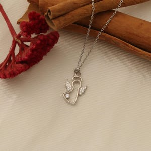 Op de afbeelding: Een zilverkleurige ketting met een engel hanger. De hanger heeft een engel omtrek met een halo en vleugels, versierd met kleine, heldere edelstenen. De ketting wordt getoond op een cr&egrave;mekleurig oppervlak met rode bloemen en kaneelstokjes.