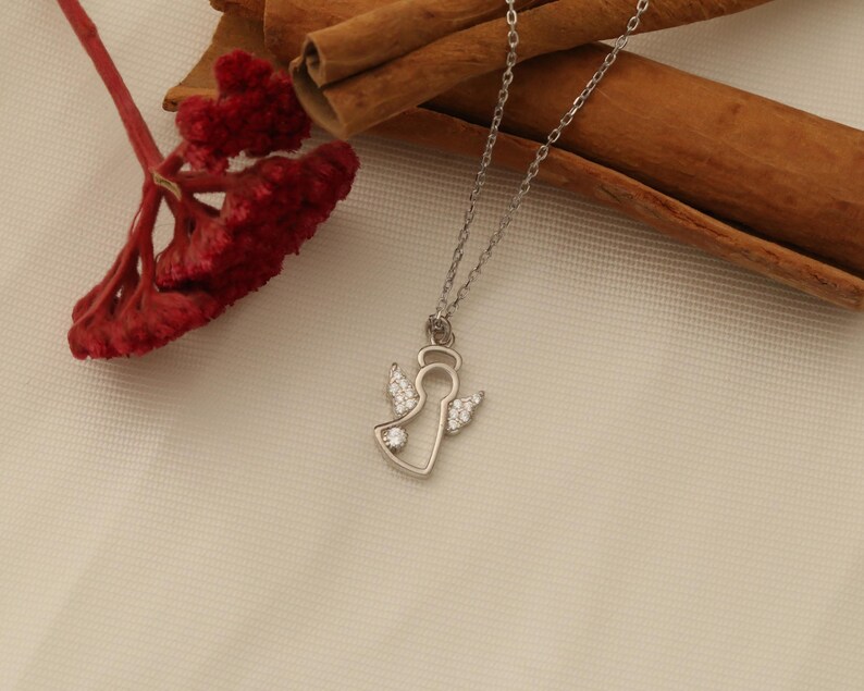 Op de afbeelding: Een zilverkleurige ketting met een engel hanger. De hanger heeft een engel omtrek met een halo en vleugels, versierd met kleine, heldere edelstenen. De ketting wordt getoond op een cr&egrave;mekleurig oppervlak met rode bloemen en kaneelstokjes.