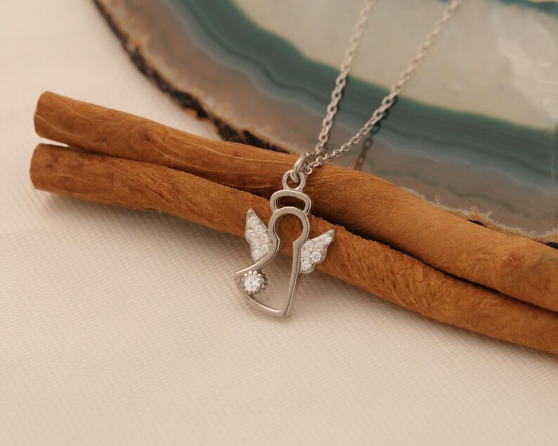 Op de afbeelding: Een zilverkleurige engel hanger ketting met een halo en met kristallen bezette vleugels. De hanger hangt aan een zilveren ketting, rustend op twee kaneelstokjes en een lichtgekleurd oppervlak.