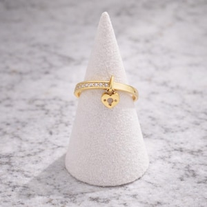 May include: A gold ring with a heart-shaped charm. The ring band is adorned with small, clear stones. The heart charm has a keyhole design. The ring is displayed on a white, cone-shaped stand.
