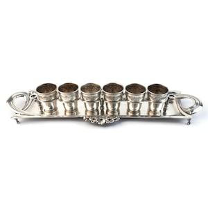 May include: A silver shot glass set with six small cups on a rectangular tray. The tray has handles and decorative floral accents. The shot glasses and tray are made of shiny silver metal.