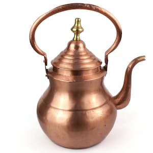 May include: A copper teapot with a curved spout and a large handle. The lid is topped with a brass finial. The teapot has a rounded body and a polished finish, reflecting the light. This decorative teapot is suitable for serving tea.